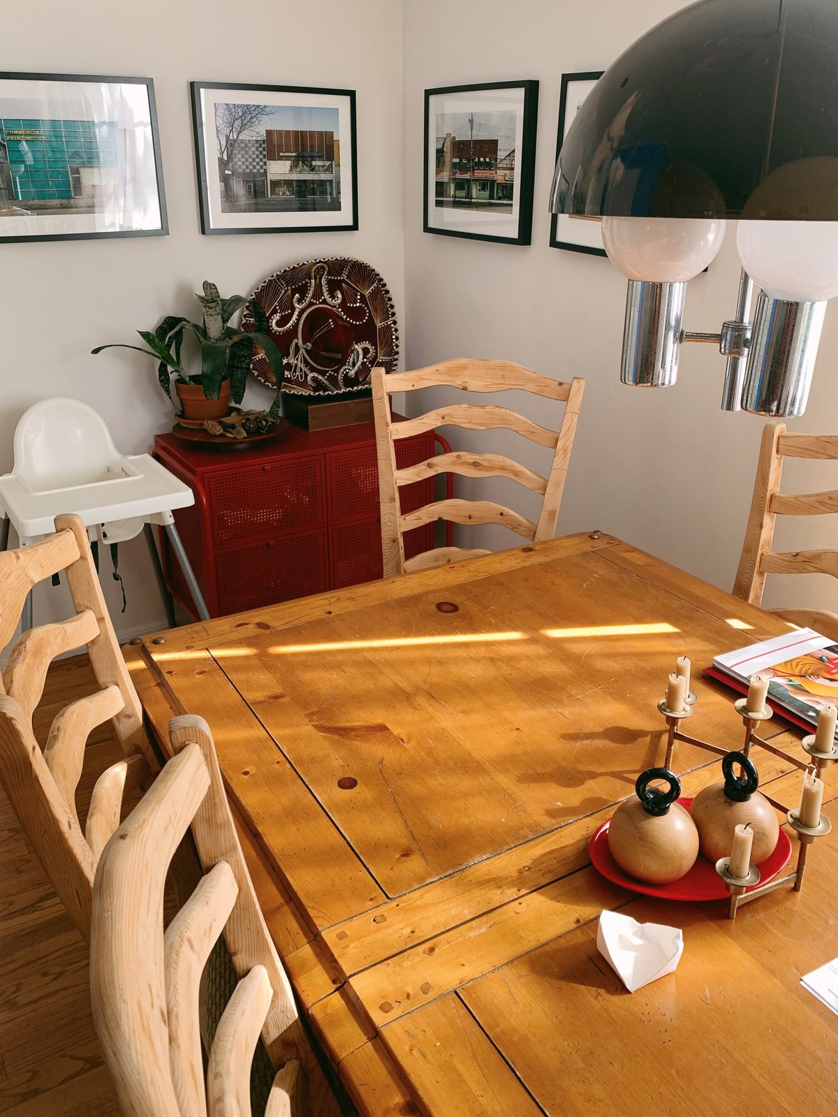 Four wooden chairs with tall backs surround a long wooden dining table. In the background is four photos displayed in black frames.