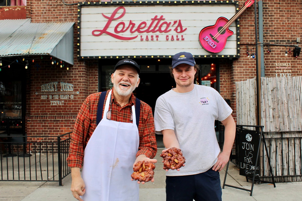 Two men stand in front of Loretta’s Last Call in Fenway, smiling and holding out donuts towards the camera.