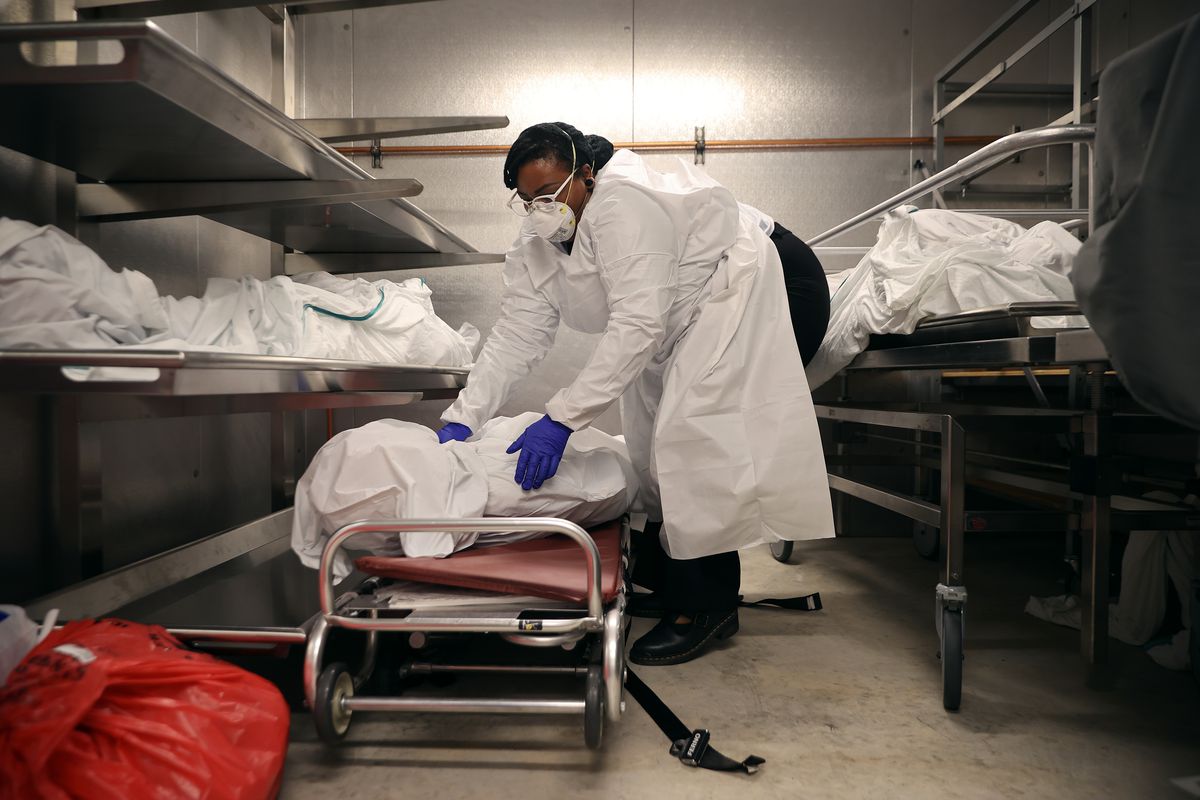 A worker wearing a mask, surgical gown, and gloves places a body wrapped in a sheet onto a stretcher inside a morgue.