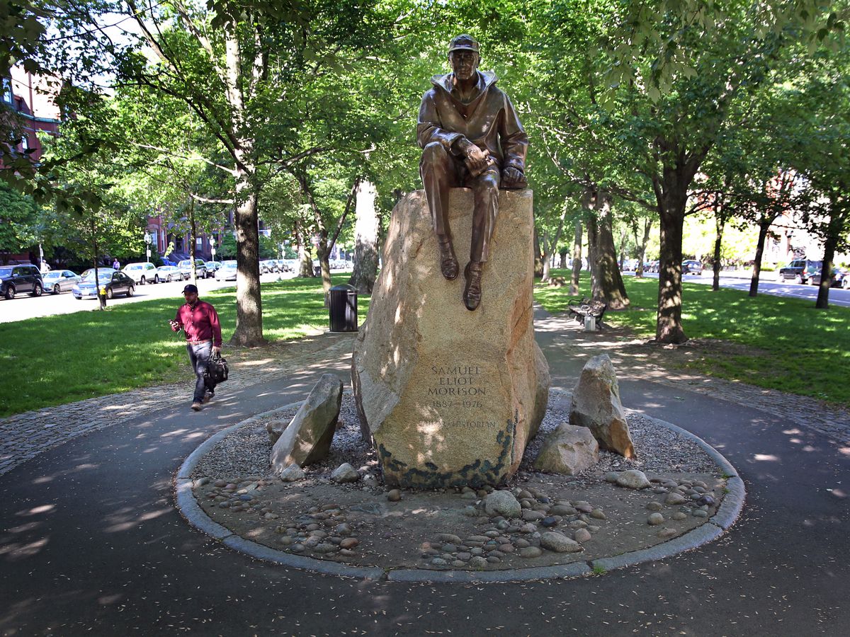 A bronze statue along a pedestrian mall. It’s of a man sitting on a big rock. 