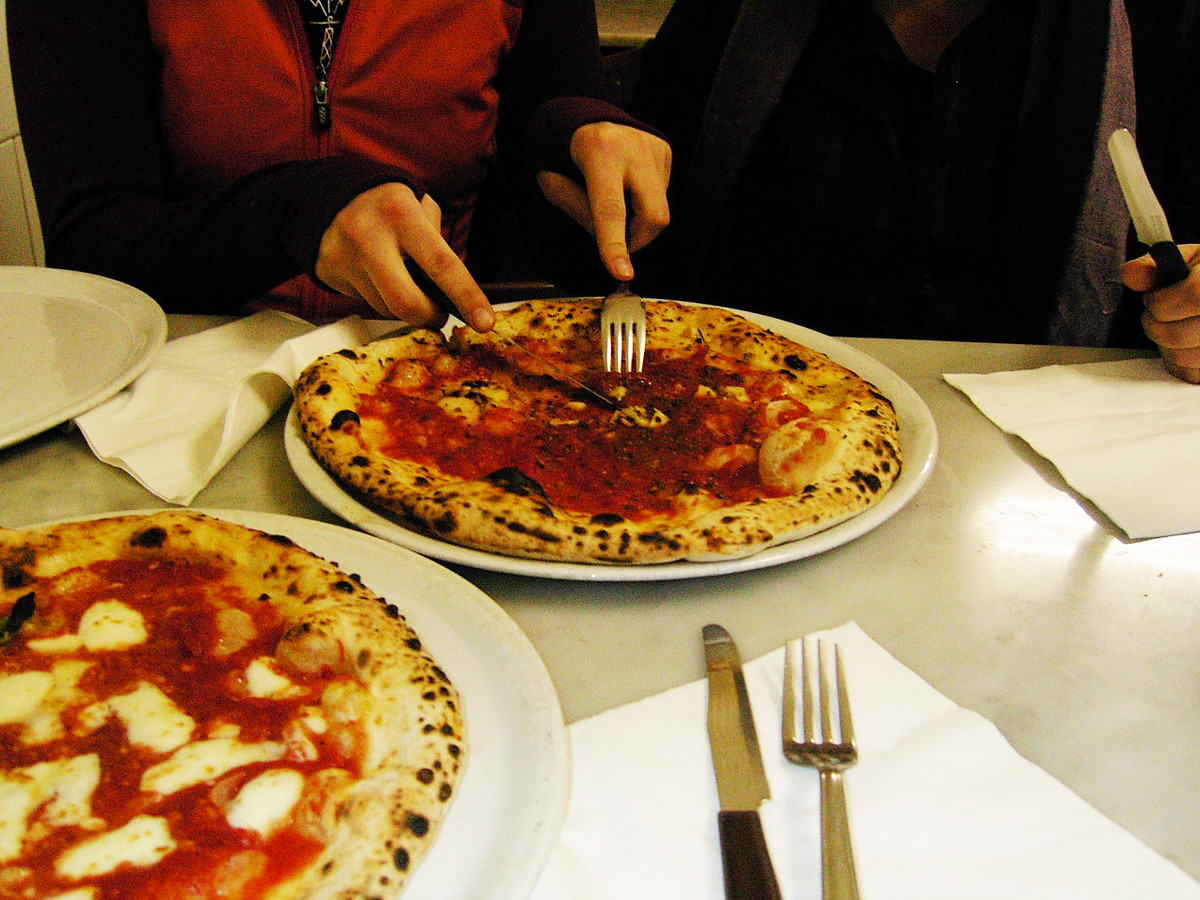 Hands holding a fork and knife cut into a whole pizza on a white plate sitting on a table. Another whole pizza is positioned on the table to the left.