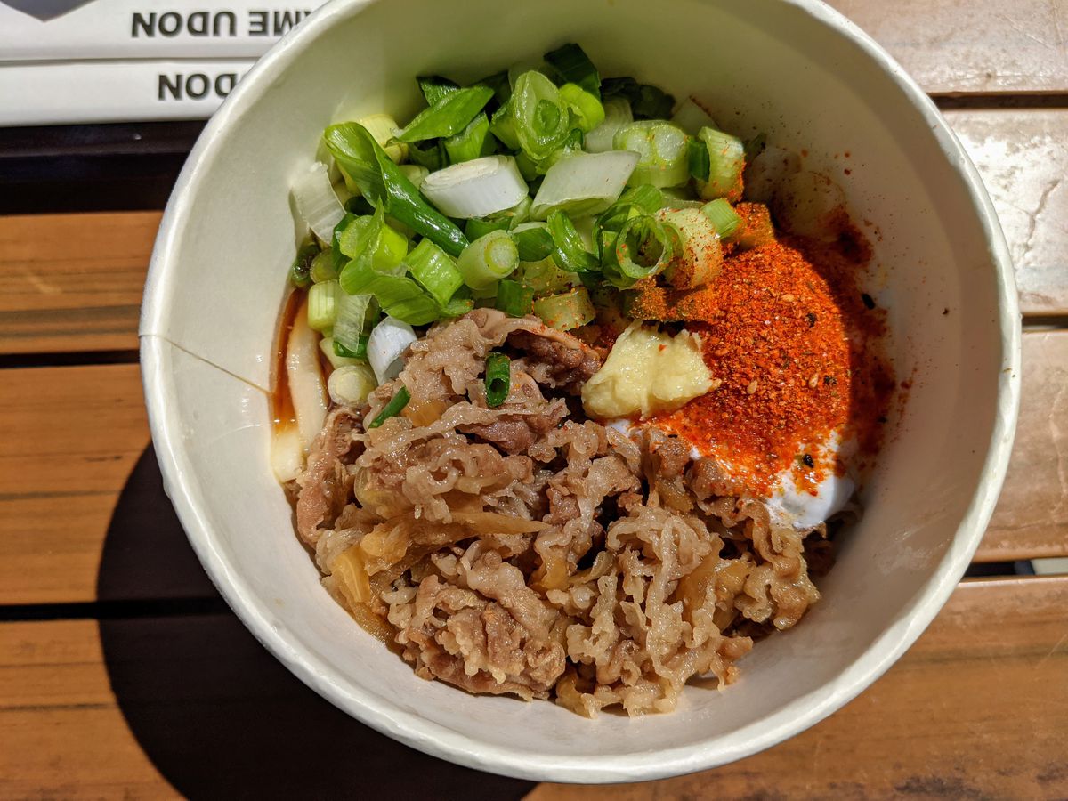 A white takeout bowl on a wood surface filled with nikutama at Marugame Udon.