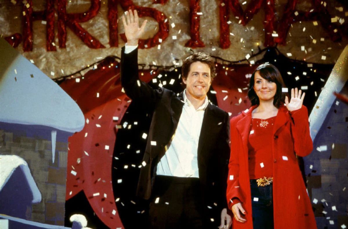 hugh grant as prime minister, waving on stage, while natalie, dressed in all red, stands awkwardly next to him