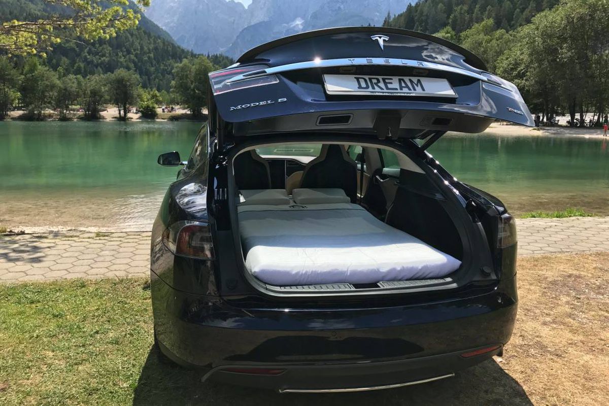 A Tesla car has its back hatch door open and a two-person bed inside. The background is a mountainous lake. 