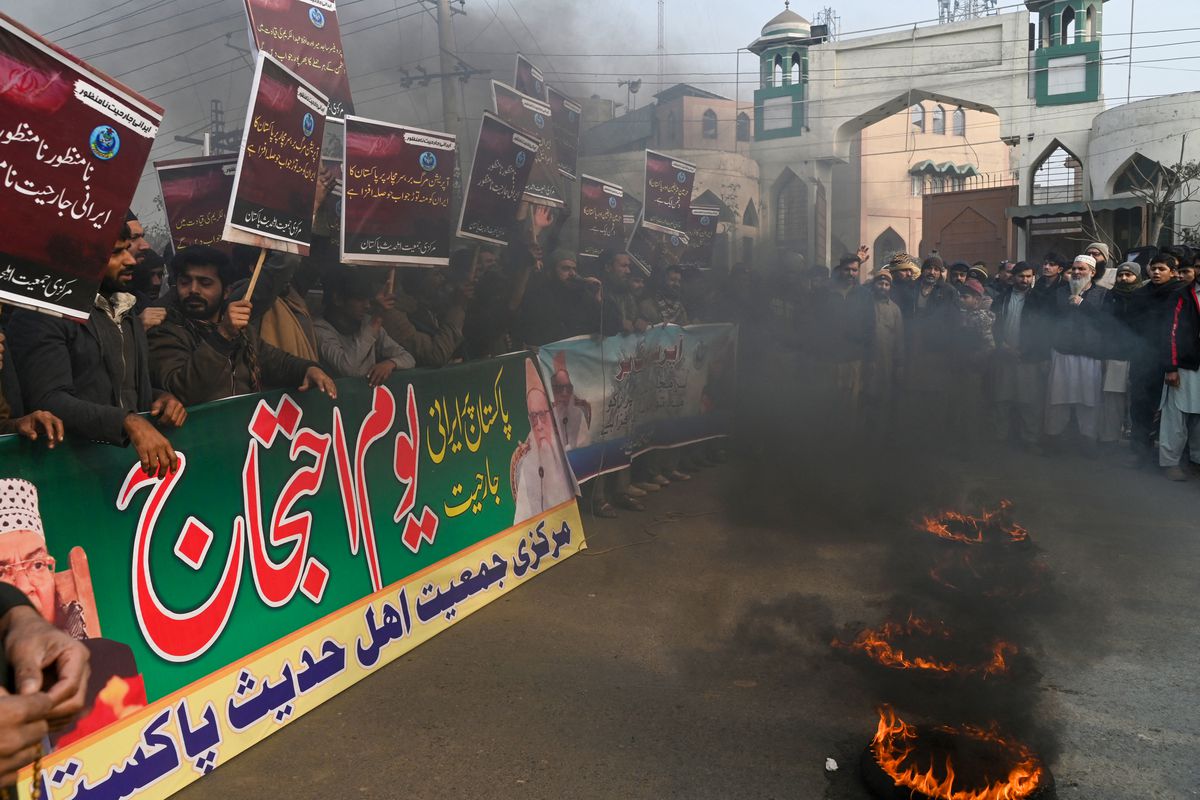 Protesters with banners and signs stand in the street, surrounded by smoke and with fire on the road in the foreground.