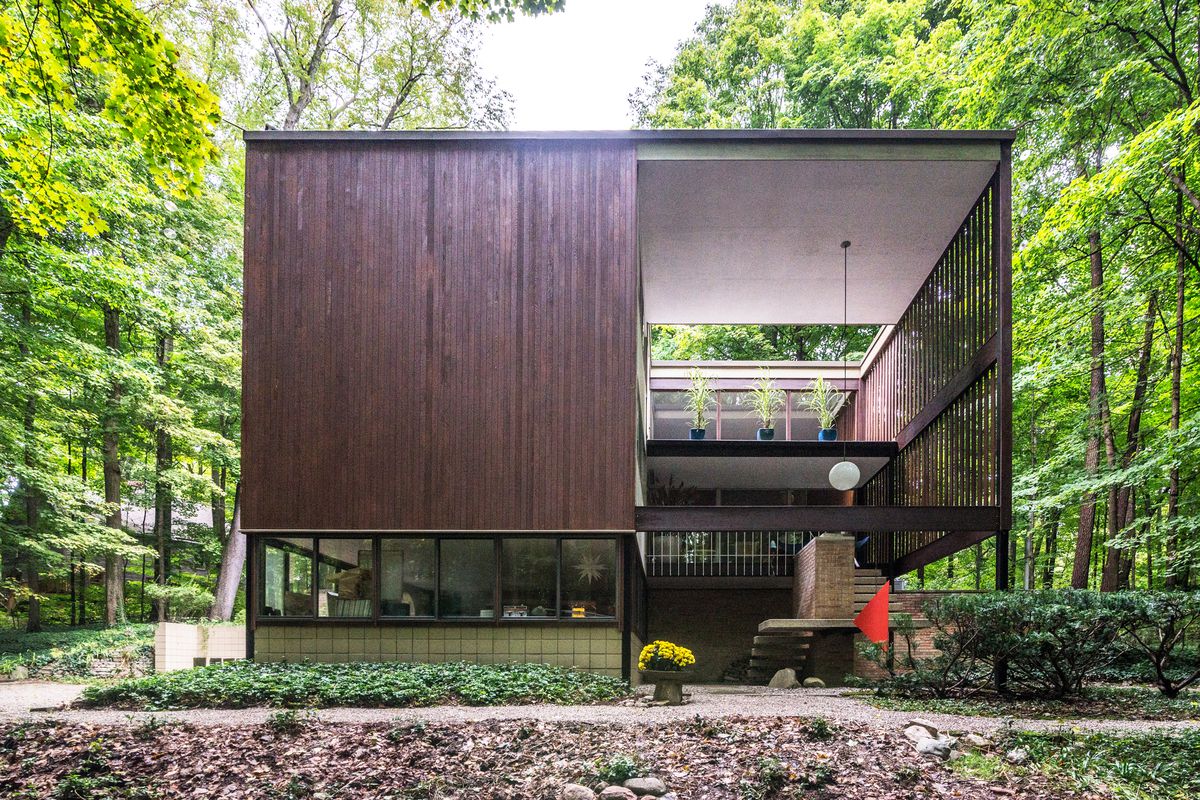 A geometric home with a solid side with wood siding and an ‘open’ side with a lofted living space. The home is surrounded by woods.