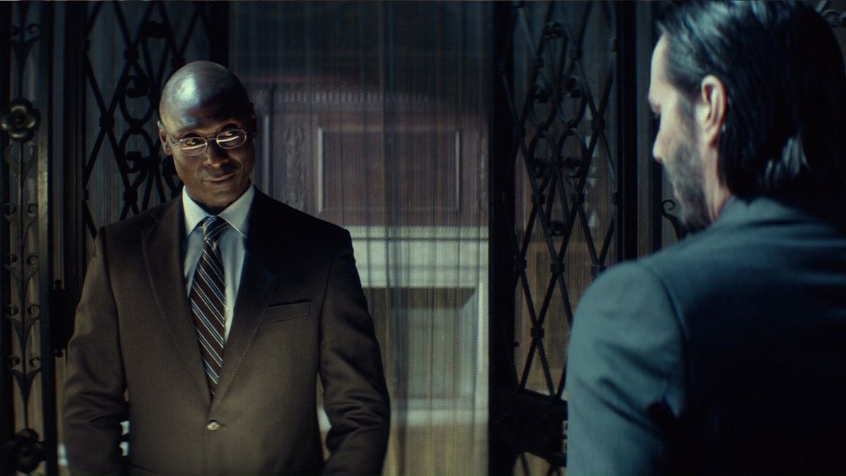 Lance Reddick as concierge Charon, in a dapper brown suit, greets Keanu Reeves as John Wick at the check-in desk of the Continental hotel in John Wick: Chapter 3 – Parabellum