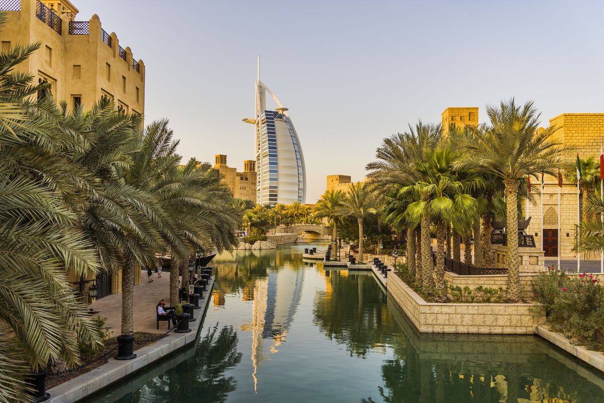 A canal lined with palm trees and stone buildings, with the Burj al Arab skyscraper in the distance filling the sky