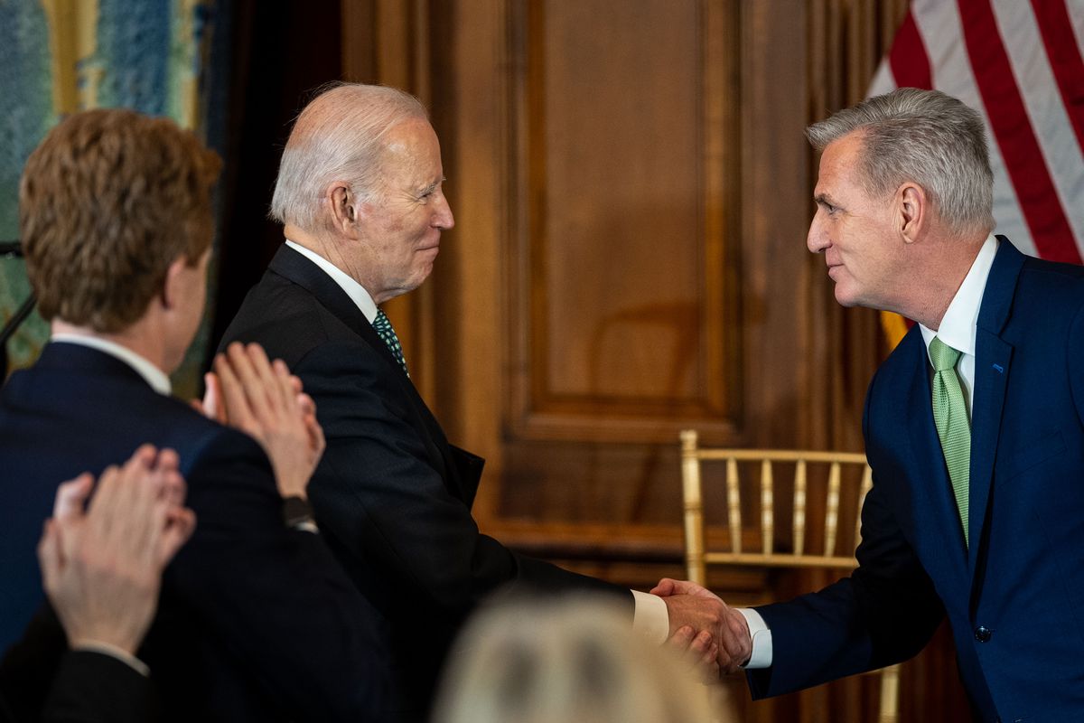 Biden and McCarthy, both smiling and besuited, shake hands in a wood paneled room as a crowd applauds.