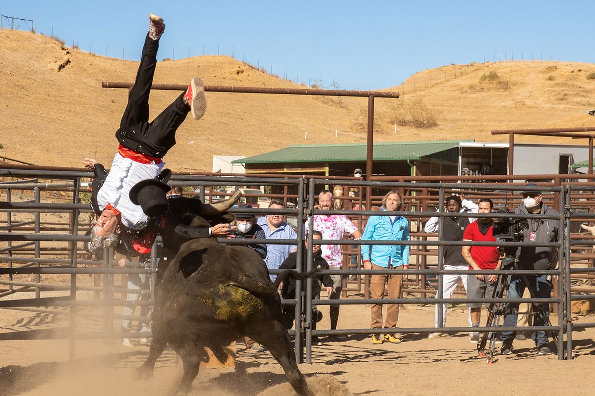 A man in a tuxedo is flying through the air upside down, having been flipped over by a bull in a ring.
