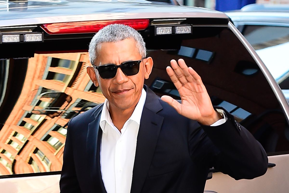 Barack Obama, wearing black sunglasses and a black suit, waves