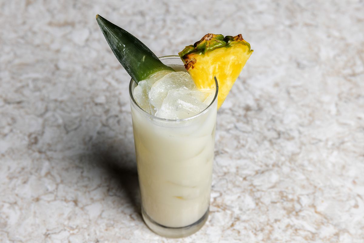 An off-white cocktail with pineapple garnish in a collins glass.