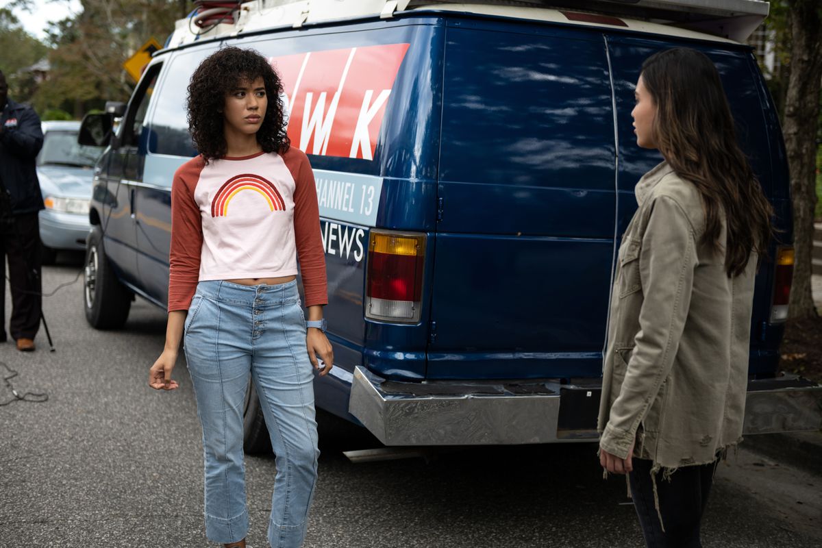 Jasmin Savoy Brown wears a long-sleeve shirt with a rainbow on it and jeans in front of a news truck in 2022’s Scream.