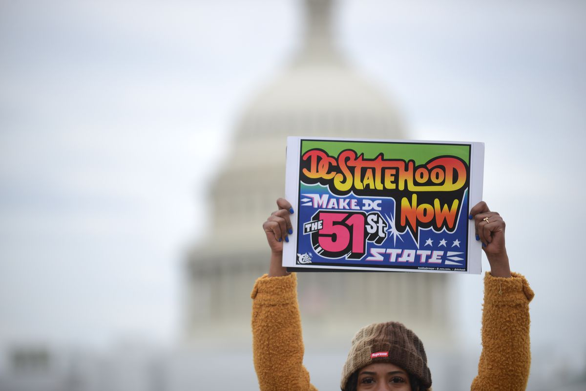 A photo showing a protestor holding a sign reading “DC STATEHOOD NOW,” with the US Capitol building blurred in the background. 