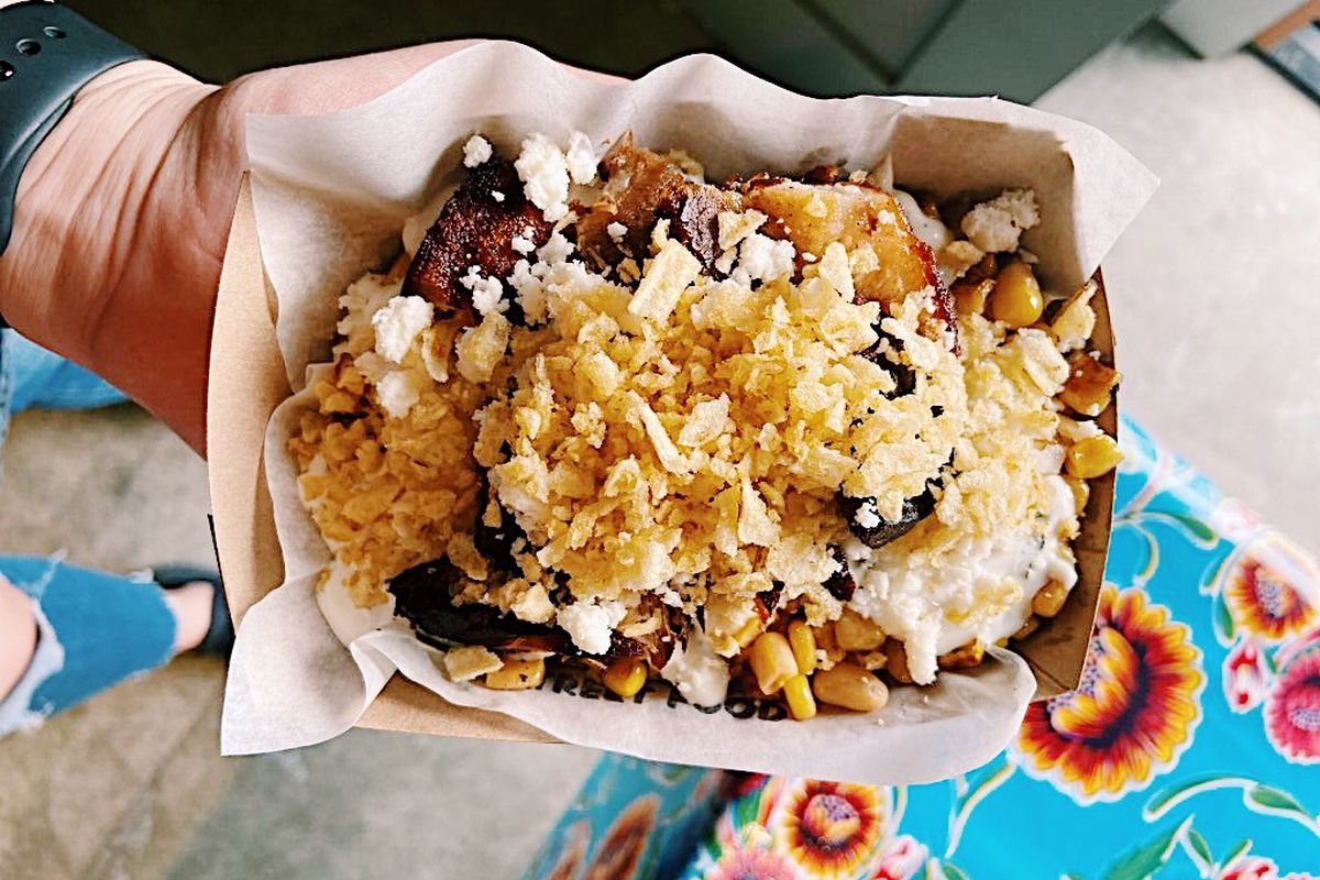 Street corn topped with crispy pork belly in a cardboard bowl with a colorful flowered tablecloth in the background