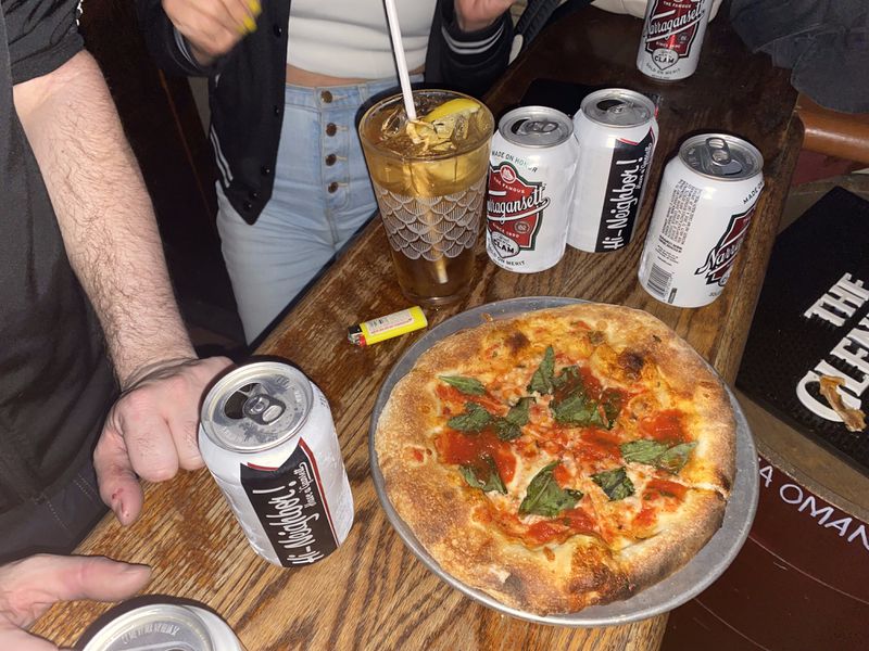 A small pizza is surrounded by cans of beer in what appears to be a dive bar.