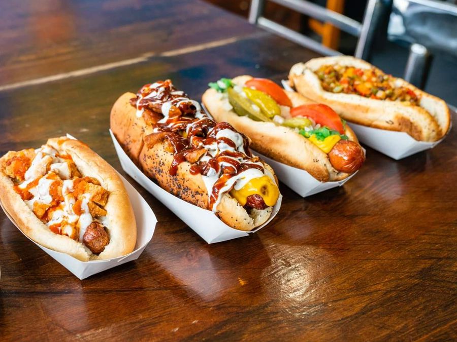 Four hot dogs with various topping sitting in white sleeves on a wooden table.