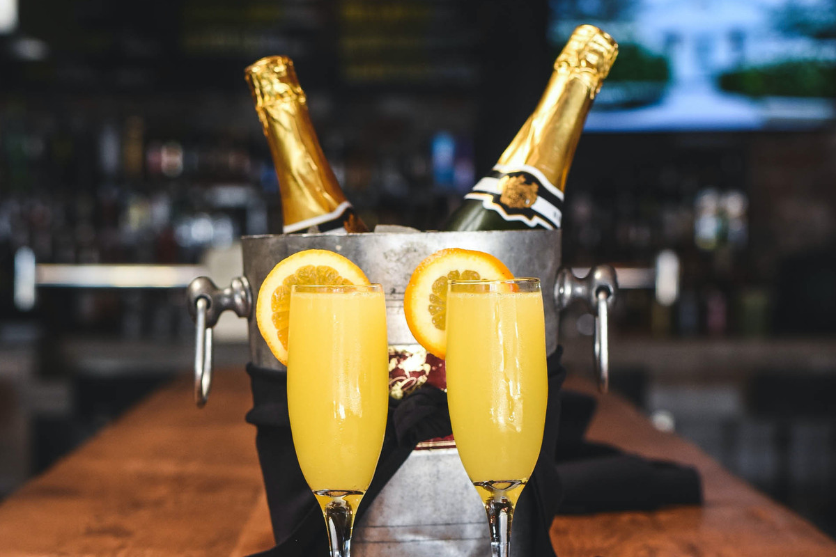 Two champagne bottles in a icy chiller flanked by mimosas with orange wedges.