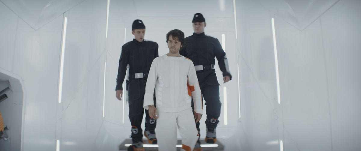 Two guards wearing grey imperial uniforms and giant insulated sneakers lead Cassian Andor, in a white body suit with orange trim, down the stairs of an all-white prison