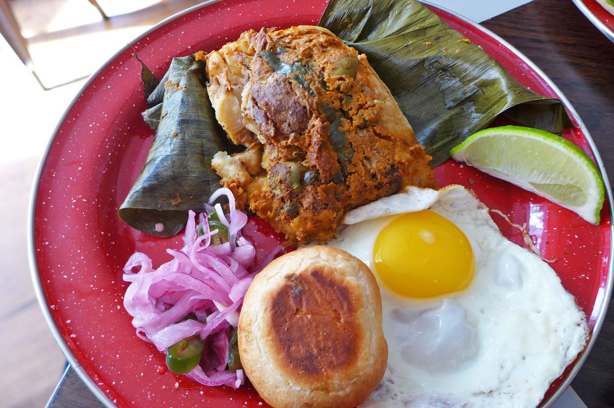 On a red plate is a banana leaf wrapped tamale, a fried egg, a roll, and pile of purple onions.