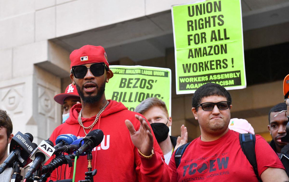 A young Black man in sunglasses and a baseball cap speaks into news outlets’ microphones, backed by supporters with signs reading, “Union rights for all Amazon workers.”