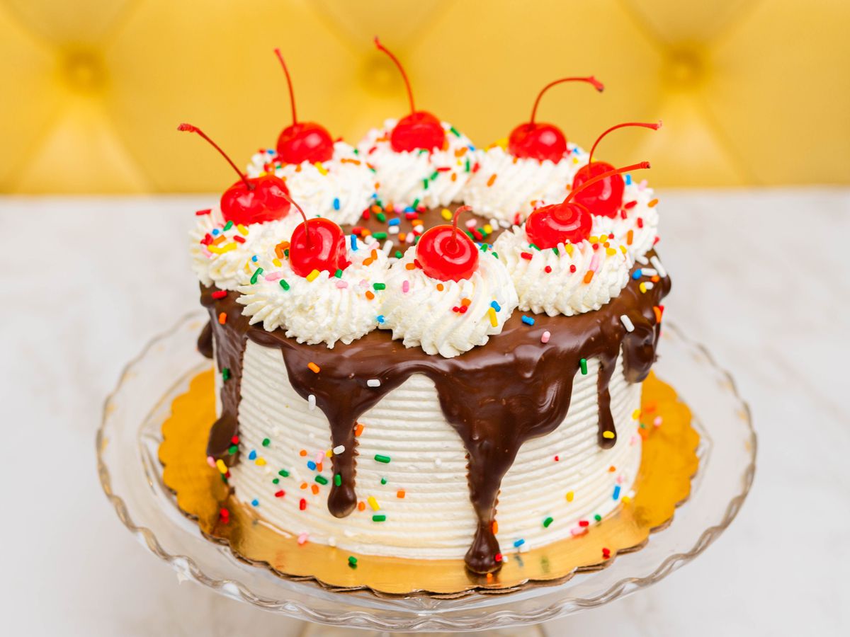 A cake frosted with white frosting with chocolate ganache on top, plus whorls of cream topped with maraschino cherries and sprinkles.
