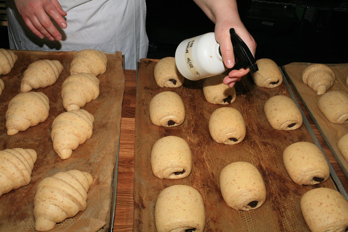 A baker sprays unbaked croissants and pains aux chocolat with egg wash.