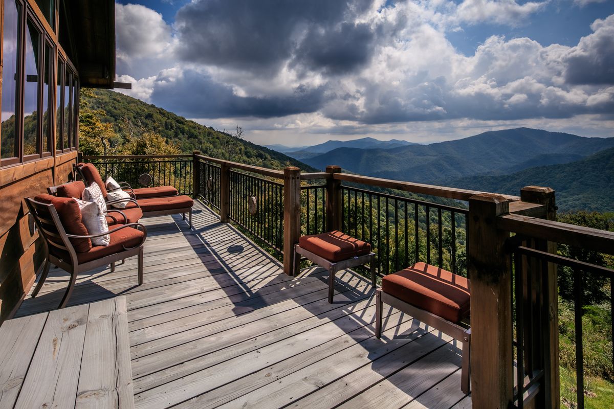 A wooden deck looks out onto rolling hills and puffy clouds in a blue sky. Red lounge furniture sits on the deck. 