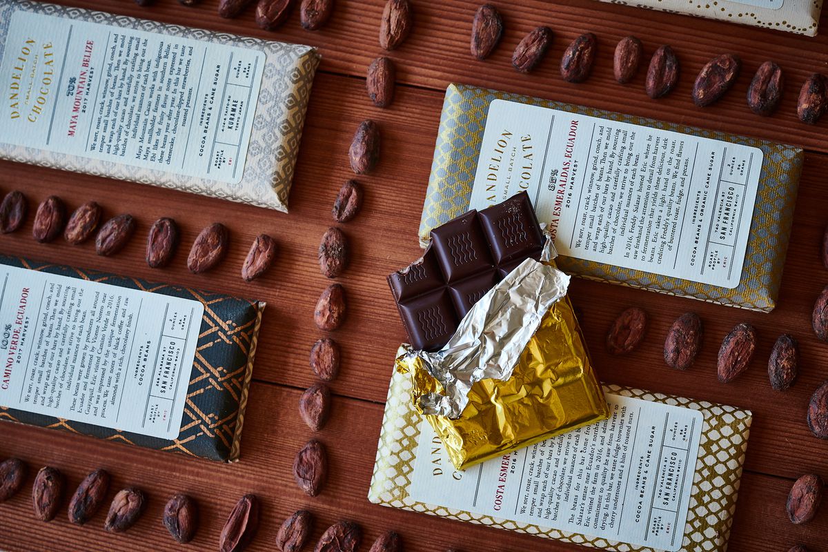Four Dandelion chocolate bars in their wrappers, in a grid with cacao beans forming lines around them.