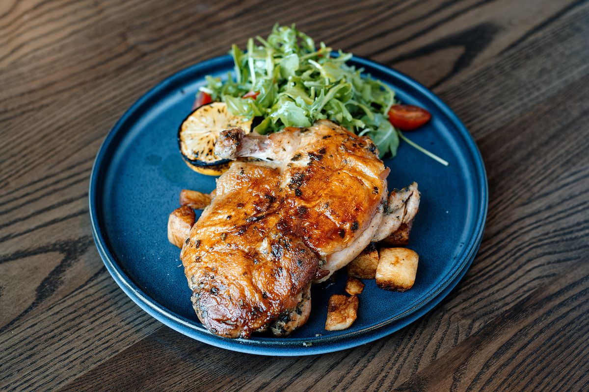 A grilled chicken spread out on a plate with greens and potatoes and a charred lemon.
