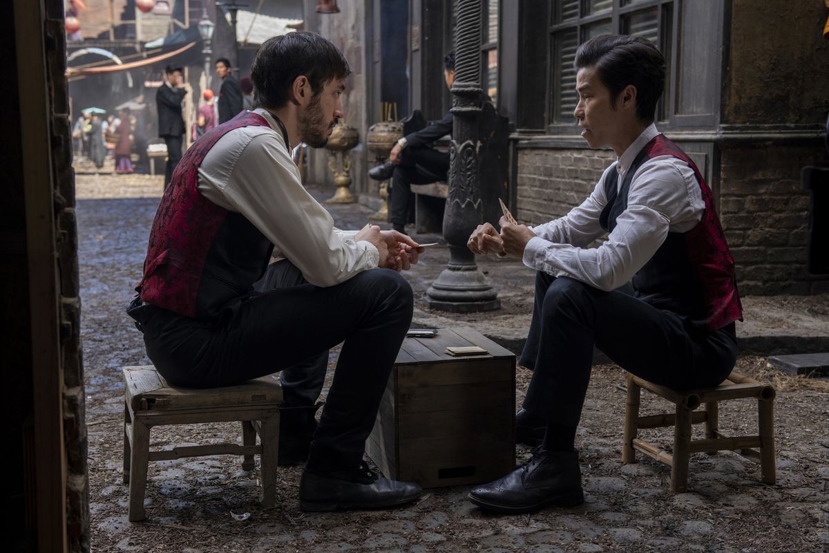 Andrew Koji and Jason Tobin, wearing red vests with white long-sleeved shirts, sit on low chairs and talk over a table in Warrior.