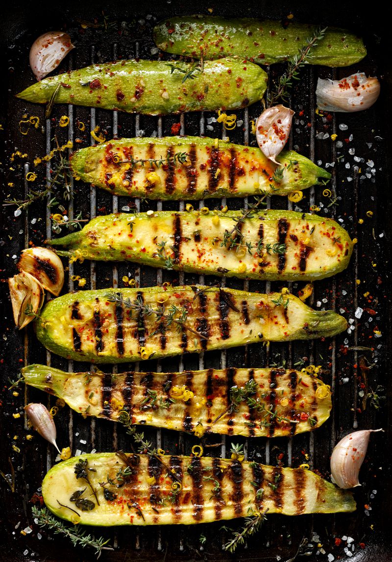 Halved zucchinis sit on a grill, with grill marks visible on the vegetables. A few cloves of garlic are scattered around them.