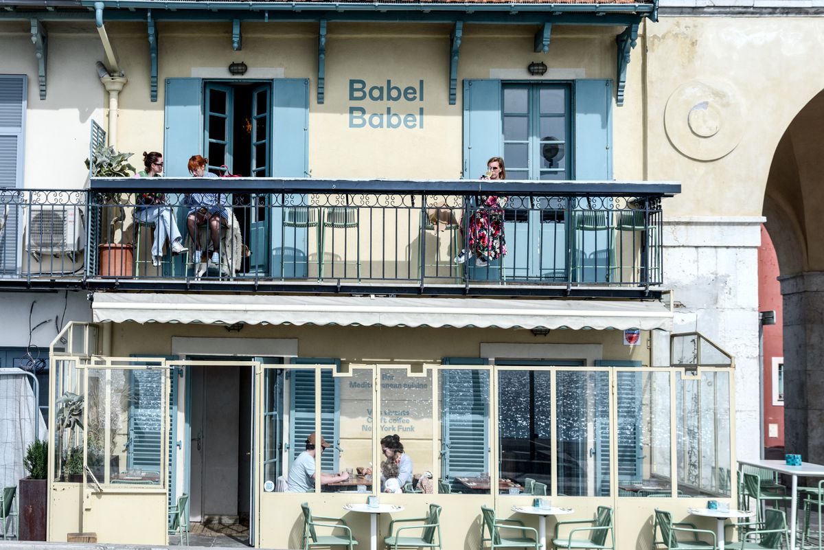 Os clientes sentam-se em mesas ao ar livre em um pátio no nível da rua e em um pátio com varanda.  O nome Babel Babel está impresso na lateral do prédio.