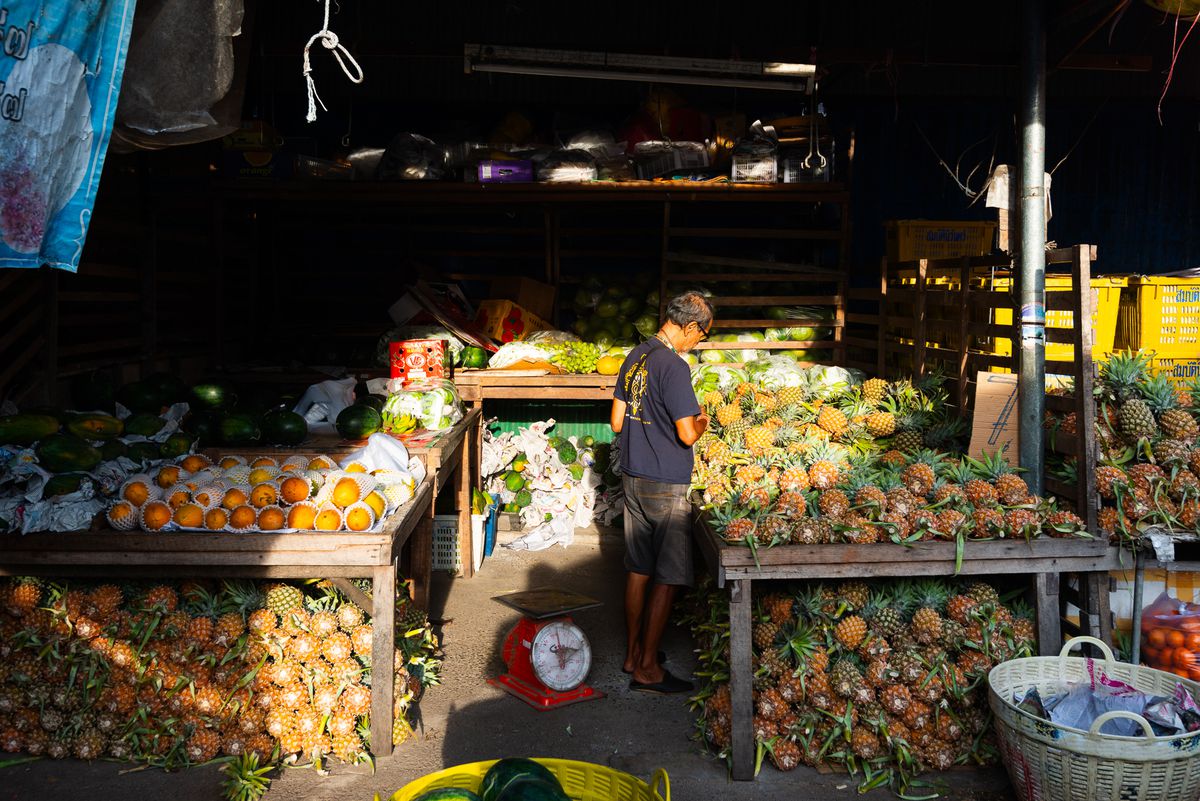 A vendor stands in a mix of sun and shadows surrounded by tables of pineapples and citrus fruits.