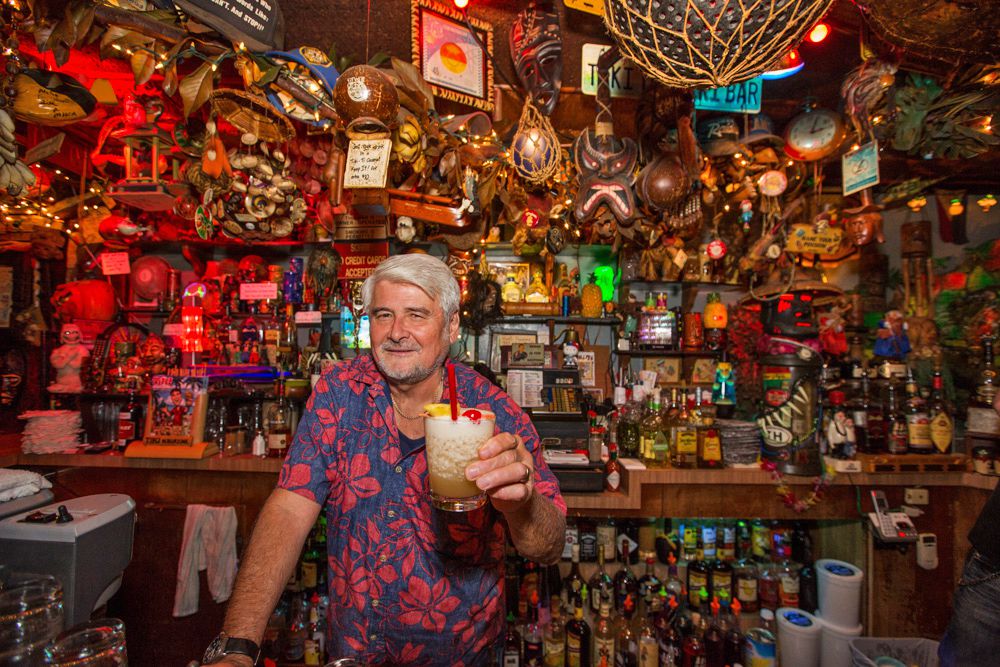 A silver-haired man in a floral shirt holds a cocktail inside a bar.