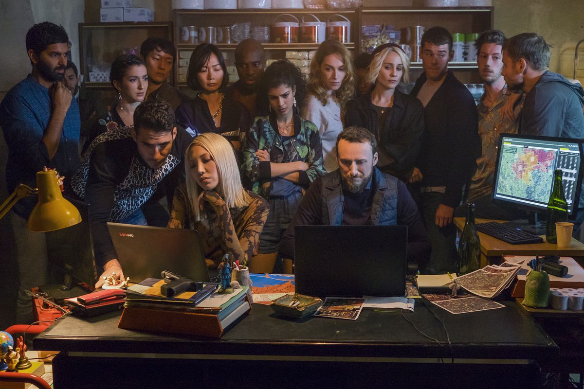 A group of people, including the principal cast of Sense8, standing over the shoulder of two people typing on laptops in Sense8.