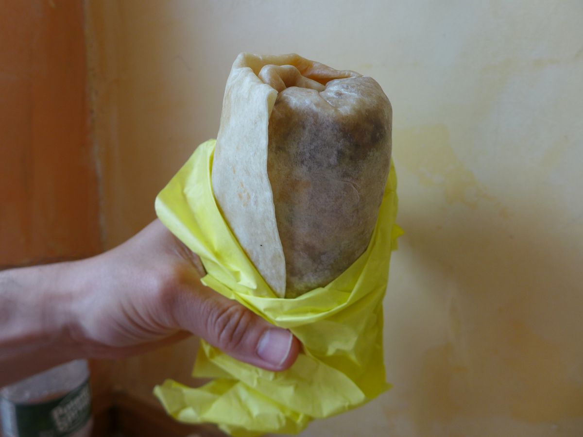 A hand holds a burrito upright in yellow tissue paper.