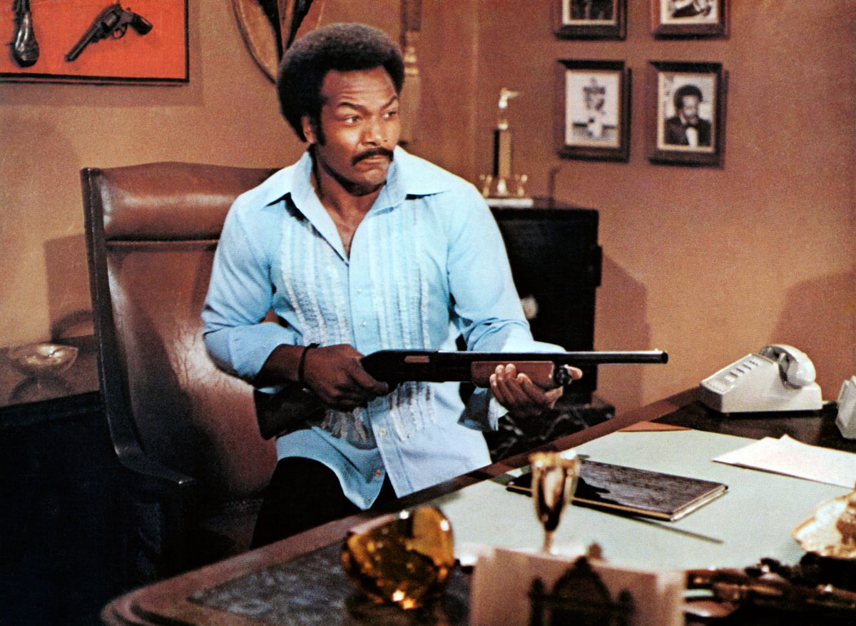 Jim Brown points a shotgun from behind a desk in Black Gunn