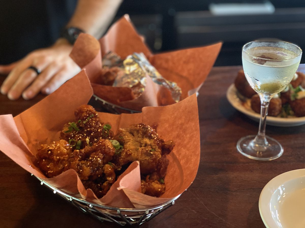 A paper-lined basked of chicken wings, another basked behind it, and a martini on a wood bar with a server standing nearby