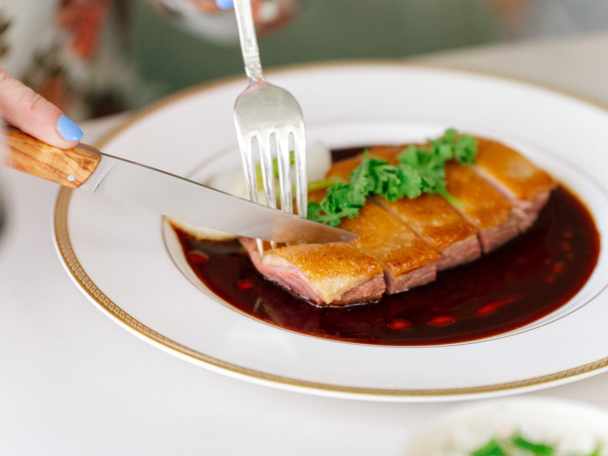 A fork and knife cutting into duck breast in foie jus on a white plate.