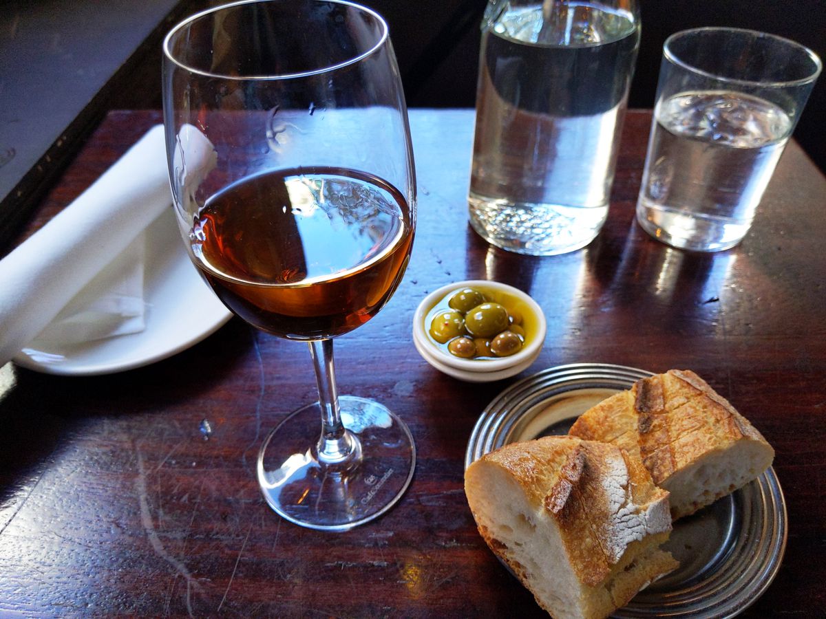 A tabletop backlit with a glass of red wine and tiny bowl of olives in oil; bread in foreground.