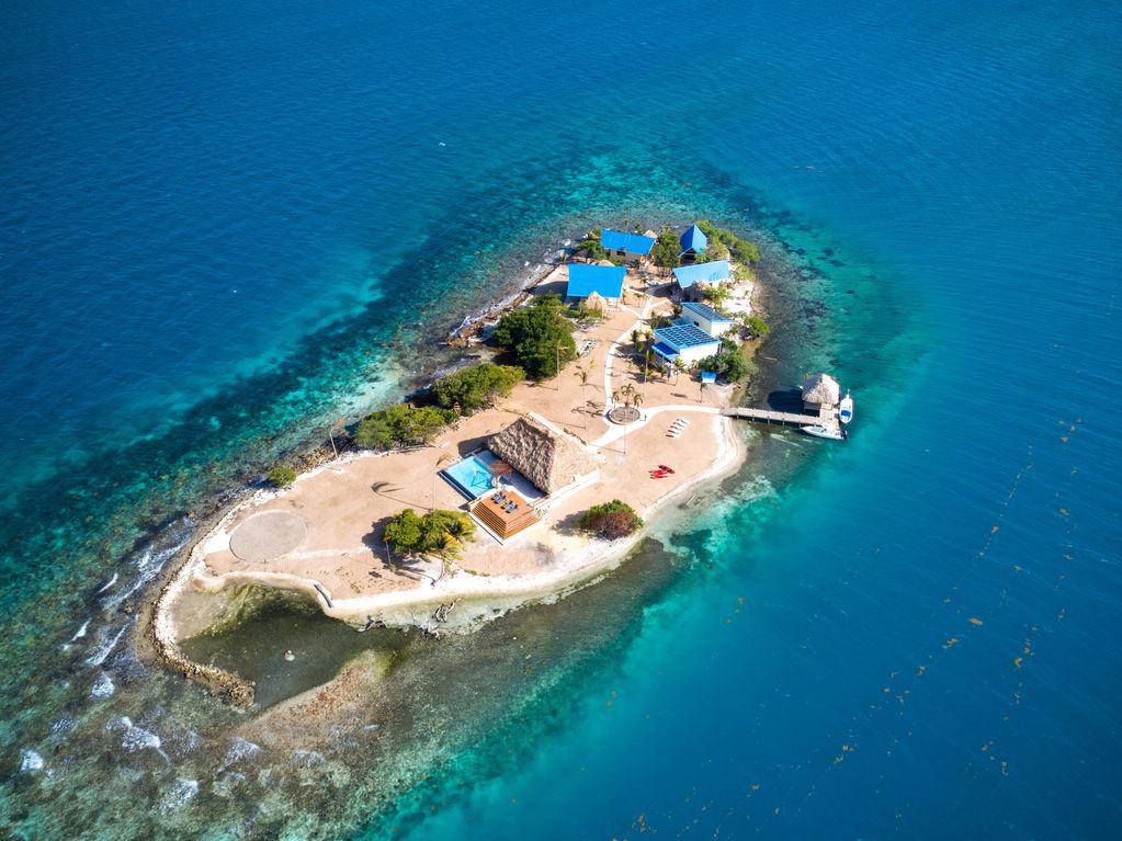 Aerial view of a private island surrounded by turquoise waters. A few buildings sit on one side of the island, where there’s also a dock.
