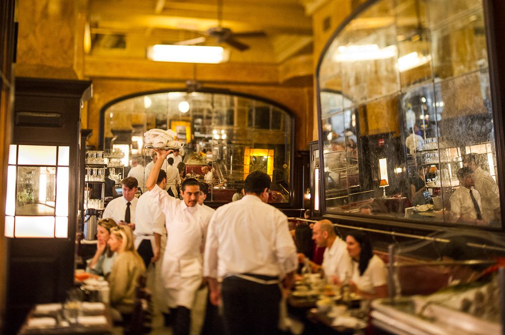Servers walk around in white in the grand Balthazar dining room, flanked in antiqued mirrors