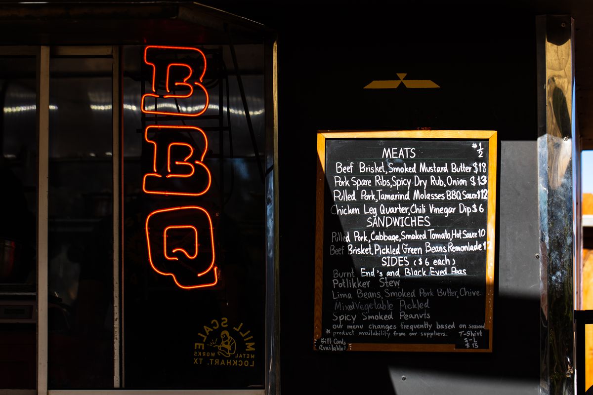 The facade of a food truck with a horizontal neon sign that reads “BBQ” next to a chalkboard side with menu items.
