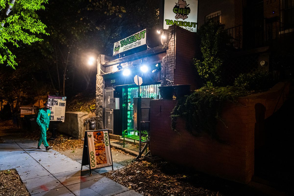 A man walks outside the green-lit front door at Chef Skip at night. 