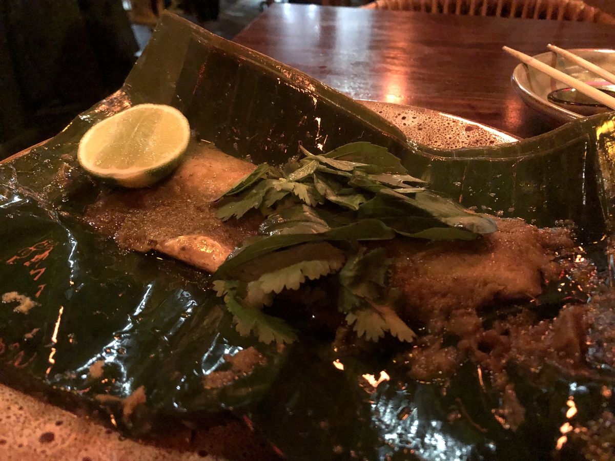A brown rectangular cut of fish set in a green banana leaf, with green herbs sprinkled over top and a lime wedge on the side.