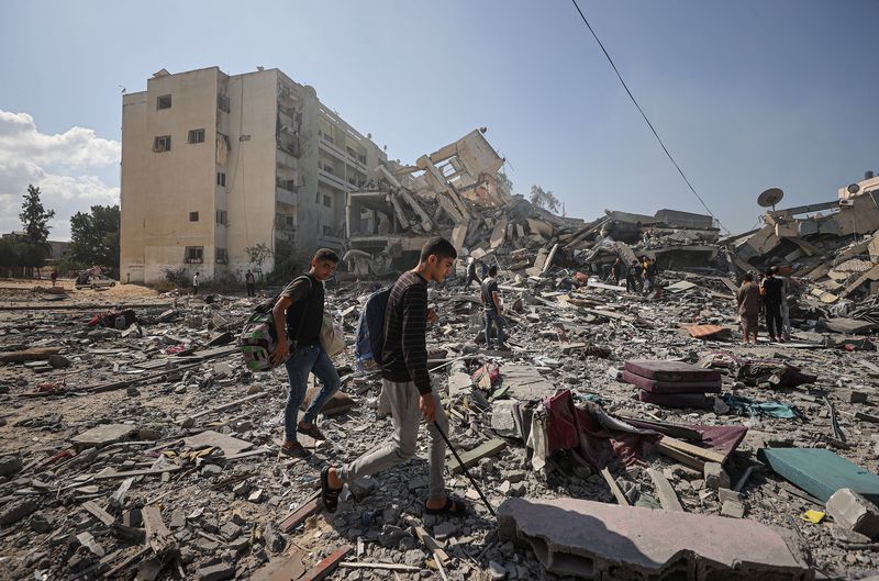 Two young Gazan men walk through rubble under a blue sky. One holds what seems to be a metal bar, and seems to use it to probe the ground. Tumbled piles of building form apocalyptic hills behind them.