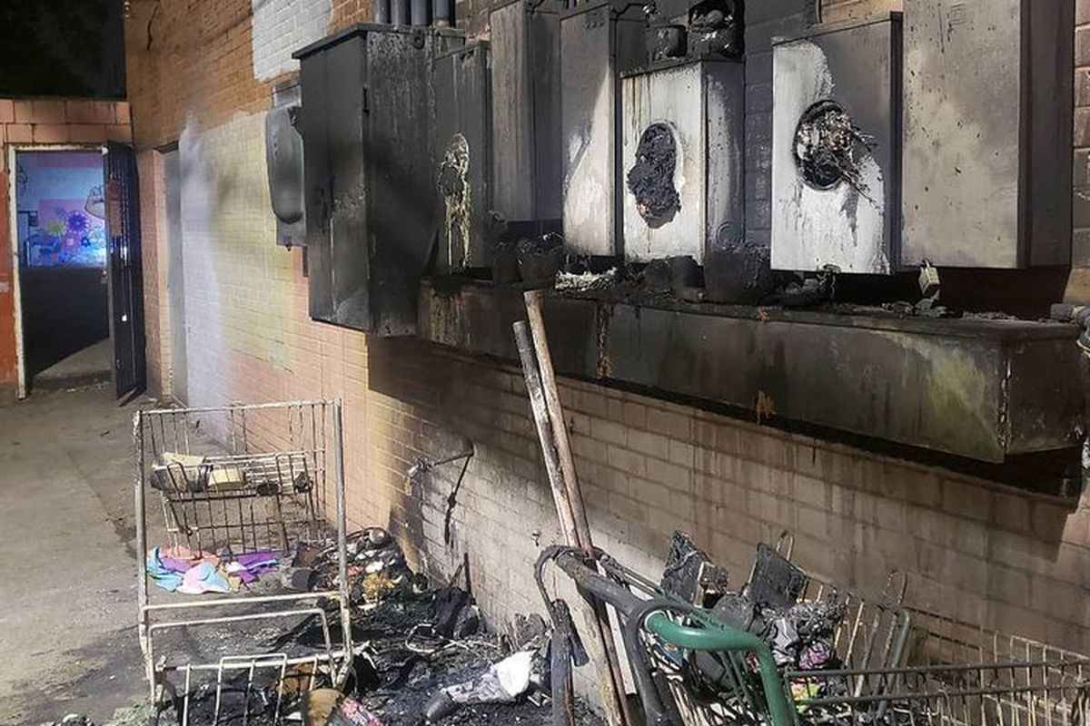 Damaged electrical panels hanging on a brick wall covered in black smoky residue from fire. Below that is two grocery store carts full of blackened burnt trash, which is also spread all over the ground. On the left, in the background is an open red door with blue light leaking out. 