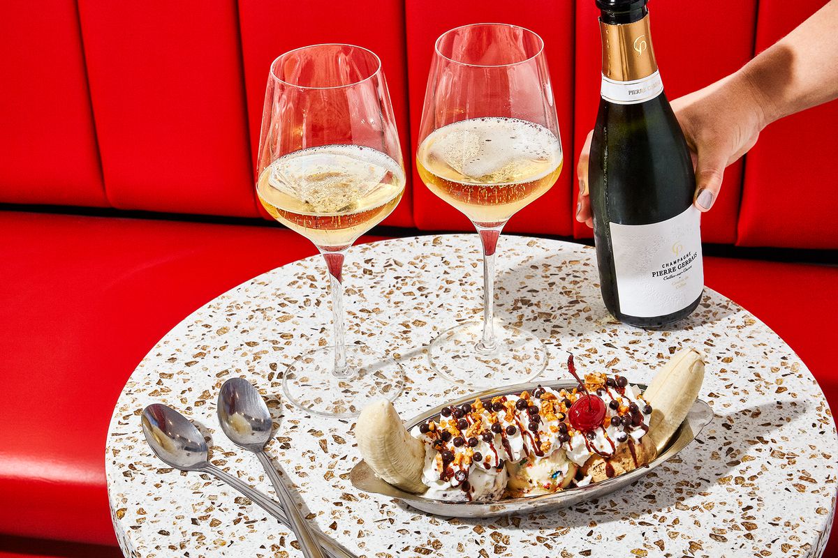 A banana split drizzled with chocolate and topped with a cherry sits on a table alongside two spoons and two glasses of champagne, against a backdrop of red banquette seating