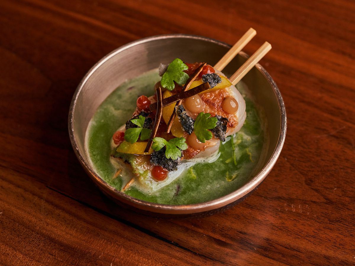 A skewered scallop with dollops of orange and red sauces over a green broth.